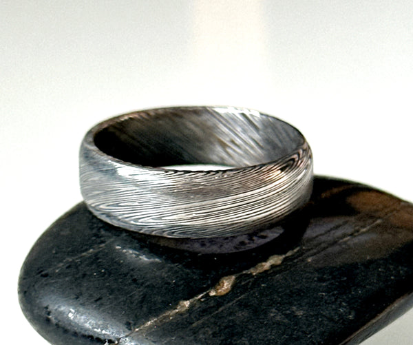 Handmade Damascus steel ring band resting on black stone, showcasing unique etched patterns.