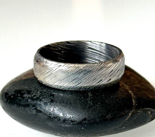 Damascus steel ring band resting on a black stone, showcasing unique etched patterns and high quality craftsmanship.