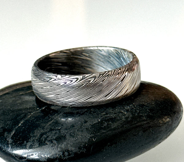 Handmade damascus steel ring band displayed on a smooth black stone, showcasing intricate etched patterns.