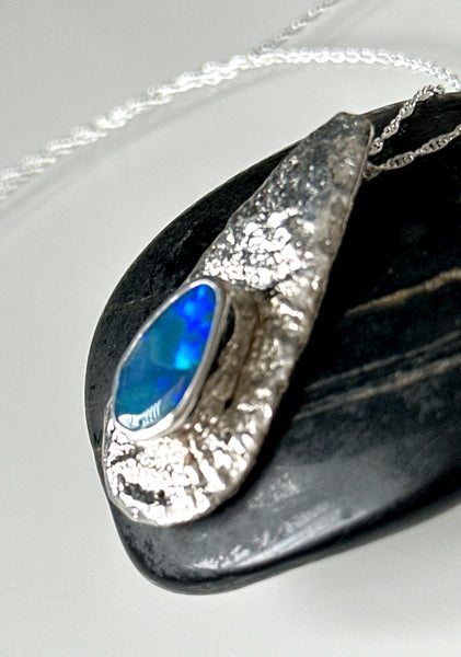 Lightning Ridge opal pendant necklace resting on a smooth black stone, showcasing vibrant blue opal in textured silver setting.